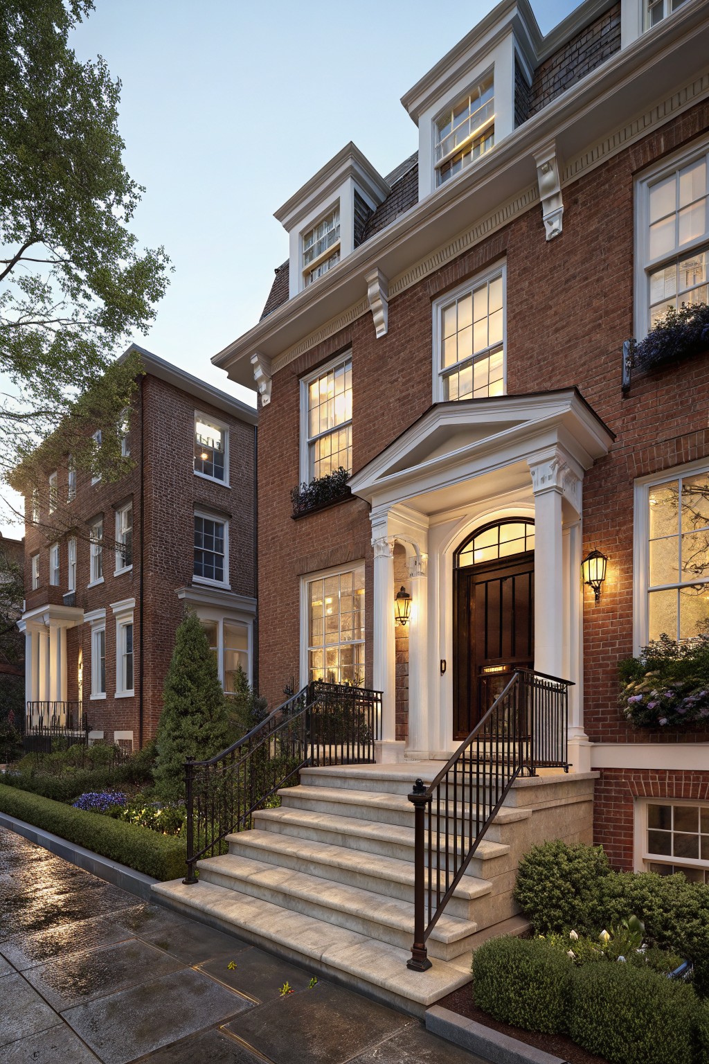 Red brick townhouse exterior at dusk with white trim, featuring a pedimented entry porch supported by columns, dark wood door, stone steps with black railings, illuminated windows, lanterns, and low shrubs along the wet street.