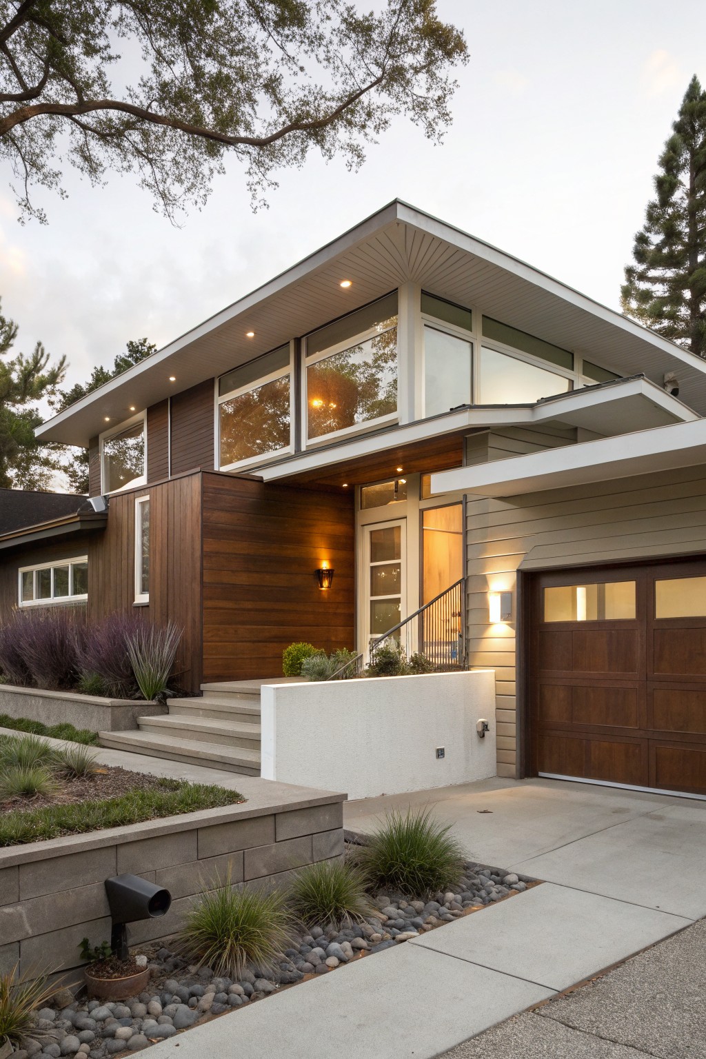Contemporary house exterior with brown wood panel cladding on entry and side walls, white roof overhangs and trim, large windows, wood garage door, concrete steps, and front yard landscaping with grasses, shrubs, rocks, and a retaining wall.