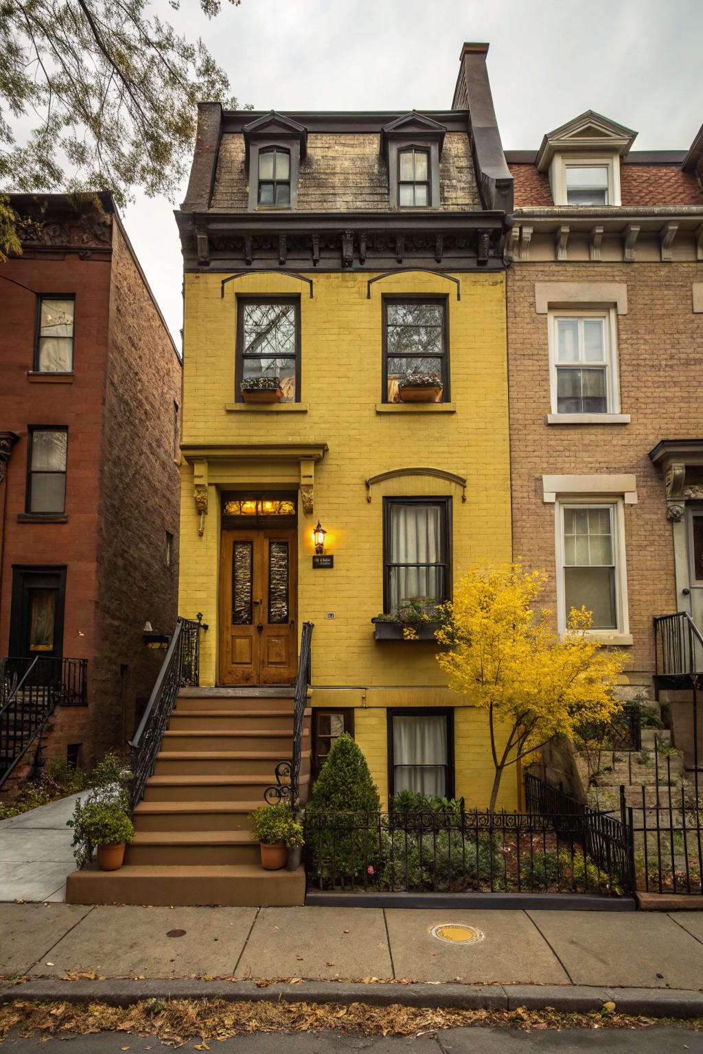 A three-story yellow brick rowhouse with brown mansard roof and dark trim, featuring a wooden front door with glass panels, lit lantern, front steps with black railing, potted plants, small tree, and garden fence, flanked by brick neighbor houses on a sidewalk.
