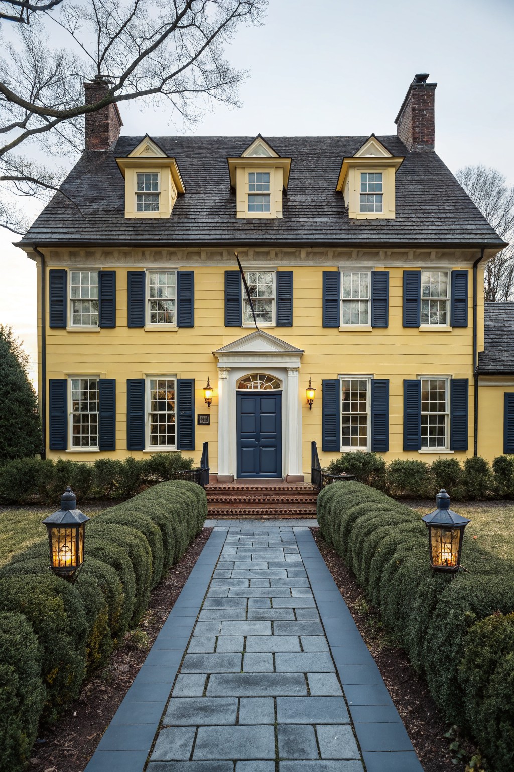 A two-story yellow clapboard house with dark shingle roof, three dormer windows, navy blue shutters on multi-pane windows, centered navy blue paneled front door with transom and sidelights, brick steps, stone walkway lined with boxwood hedges, and lanterns on either side.