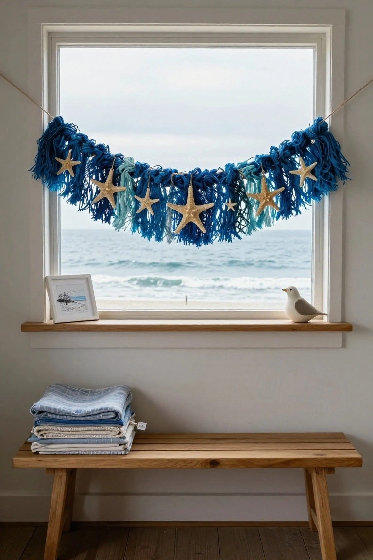 Window overlooking the ocean with a blue and teal tassel garland strung across it and decorated with starfish, a framed beach drawing and seagull figurine on the sill, and folded blankets on a wooden bench below.