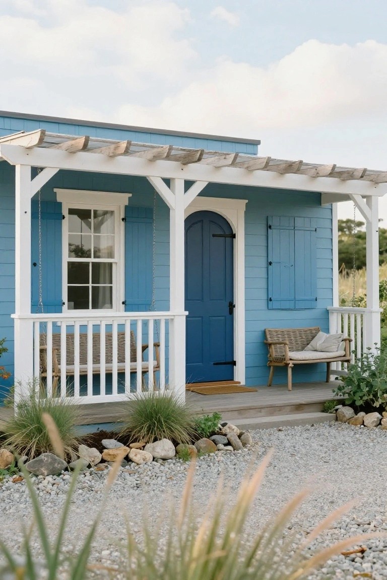 Small blue shingled cottage with white porch posts, railing, hanging swing, wicker bench, arched blue door, louvered shutters, and gravel yard edged with grasses and rocks.