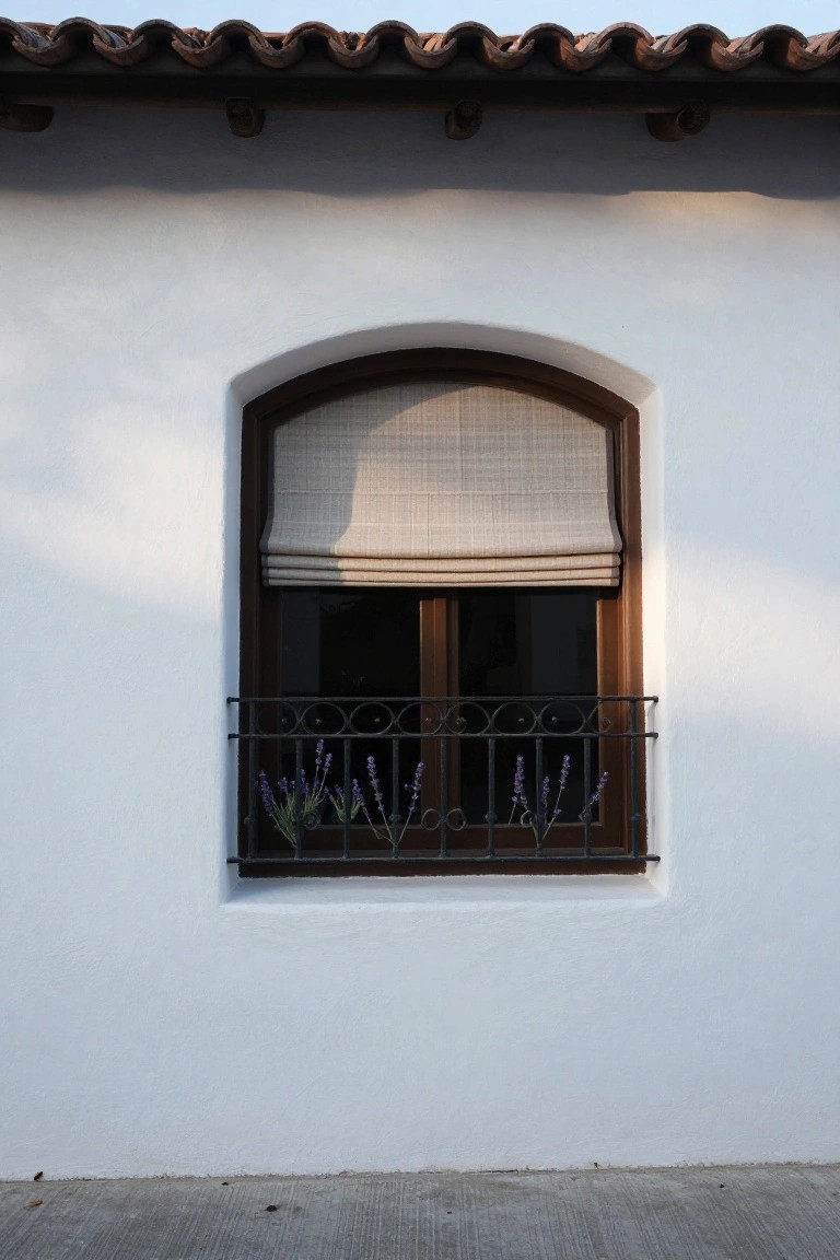 Arched exterior window with beige bamboo roman shade, dark wooden frame, black wrought iron balcony railing with lavender plants, set in white stucco wall under terracotta tile roof.