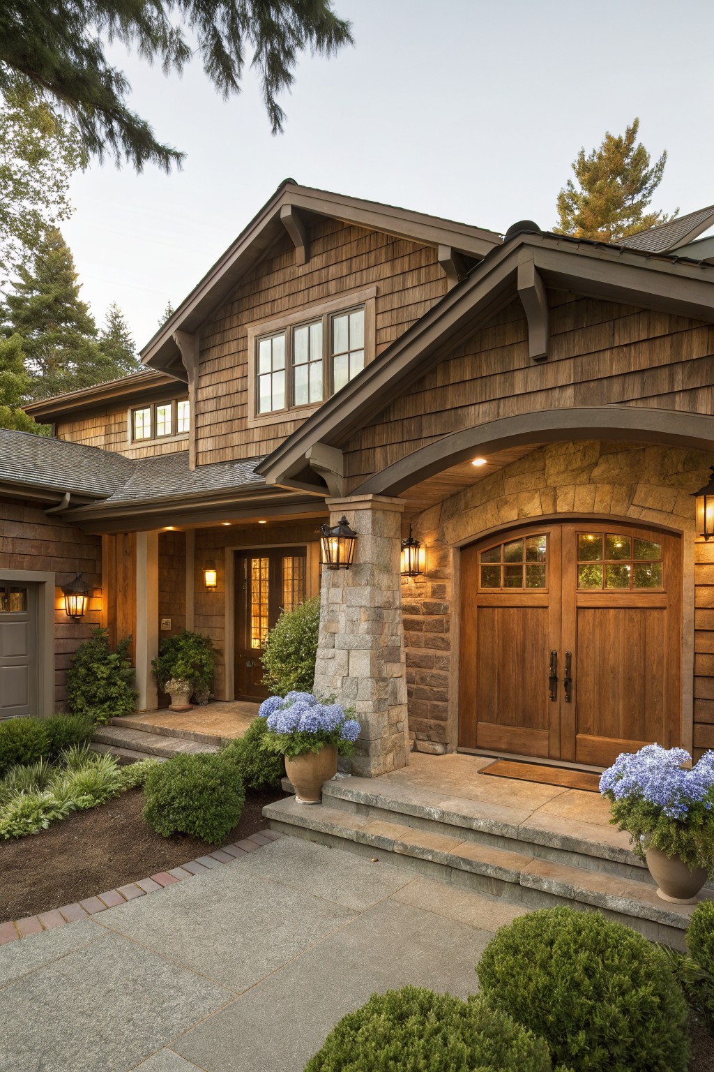 Brown shingle-sided house exterior with arched stone garage doors, wooden entry beside it, lanterns, potted blue hydrangeas, boxwoods, and stone steps leading to a concrete walkway.