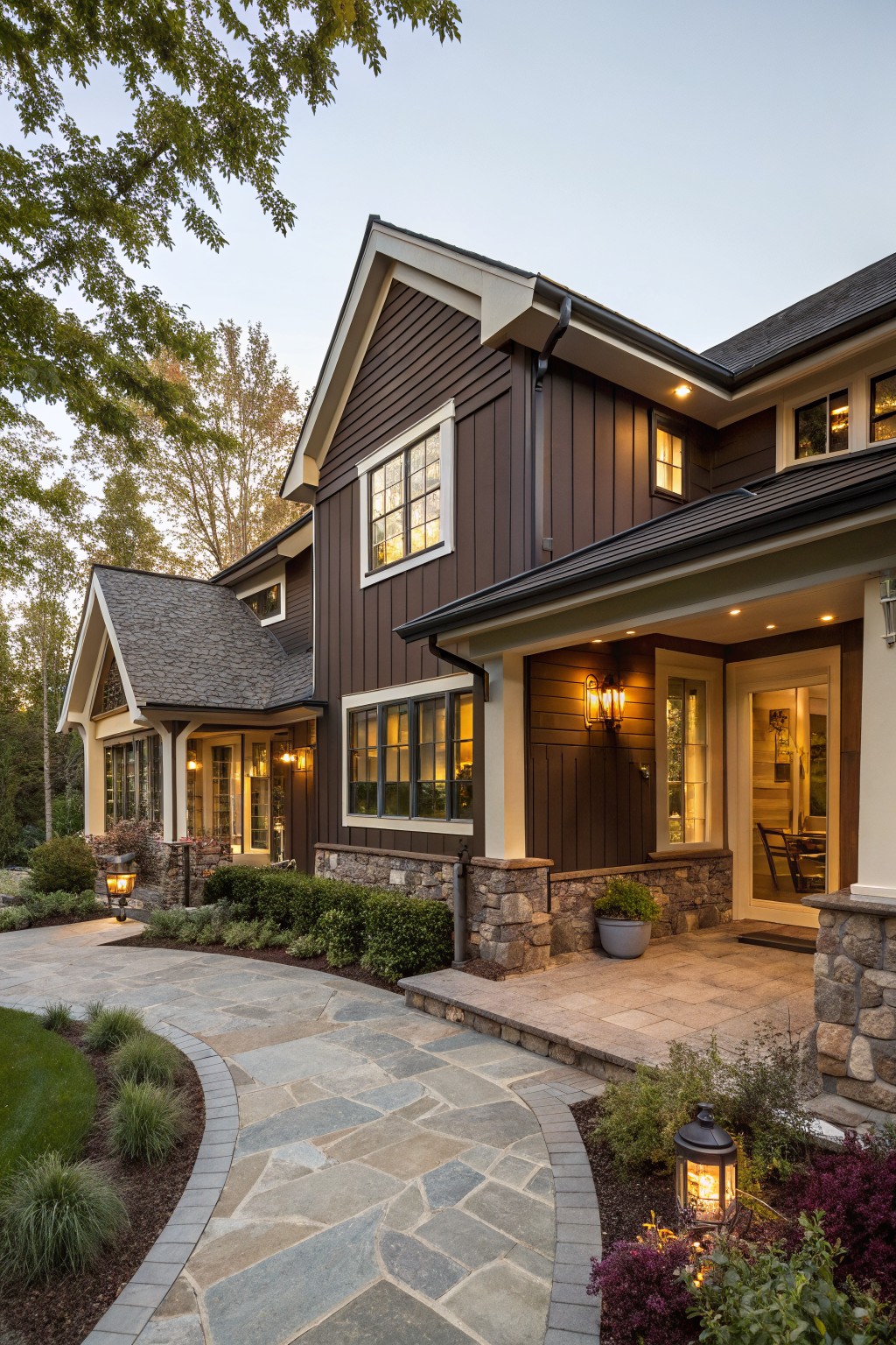 Dusk view of a brown vertical-sided house with gabled roof sections, white trim, stone foundation details, lit windows and porch lanterns, and a curved flagstone pathway through landscaping to the front entry.