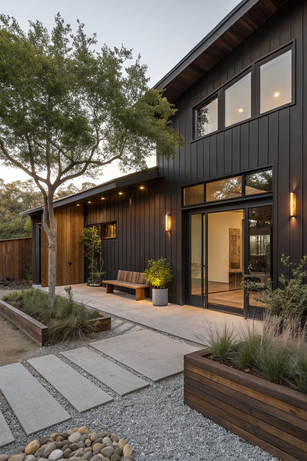 Ranch house exterior with dark vertical wood cladding, large sliding glass doors and windows, wooden bench near entry, stone pathway, raised planters with grasses, and gravel ground cover.