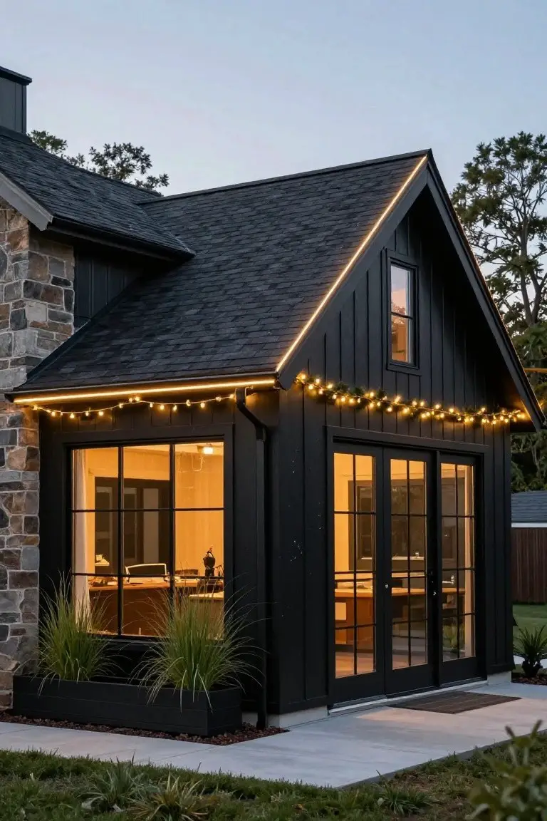 Black board-and-batten house addition with stone foundation, large black-framed windows, LED strip lights outlining the roof edges, string lights along the eaves, and potted grasses in front at dusk.
