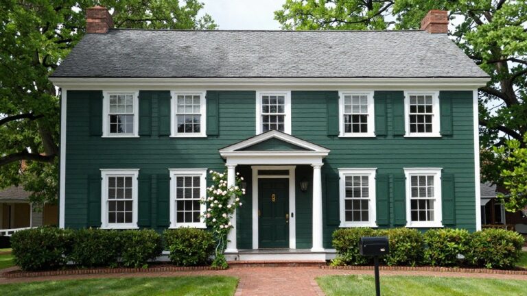 20 Dark Green Houses With White Trim That Pop Beautifully Front view of a two-story dark green clapboard house with white trim, columned porch, dark green front door, white shutters on multipane windows, flanked by shrubs, and a brick walkway.