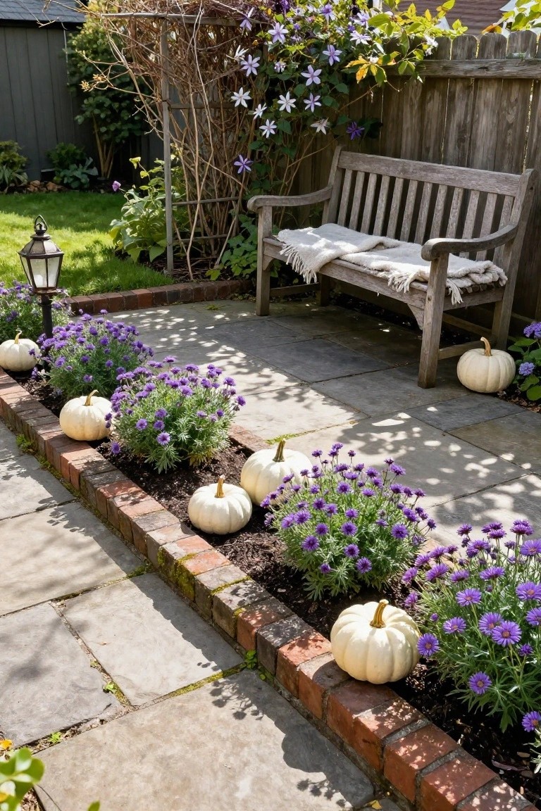 Stone patio edged with brick planters of purple flowers and white pumpkins, wooden bench with blanket nearby, garden lantern and climbing plants in background