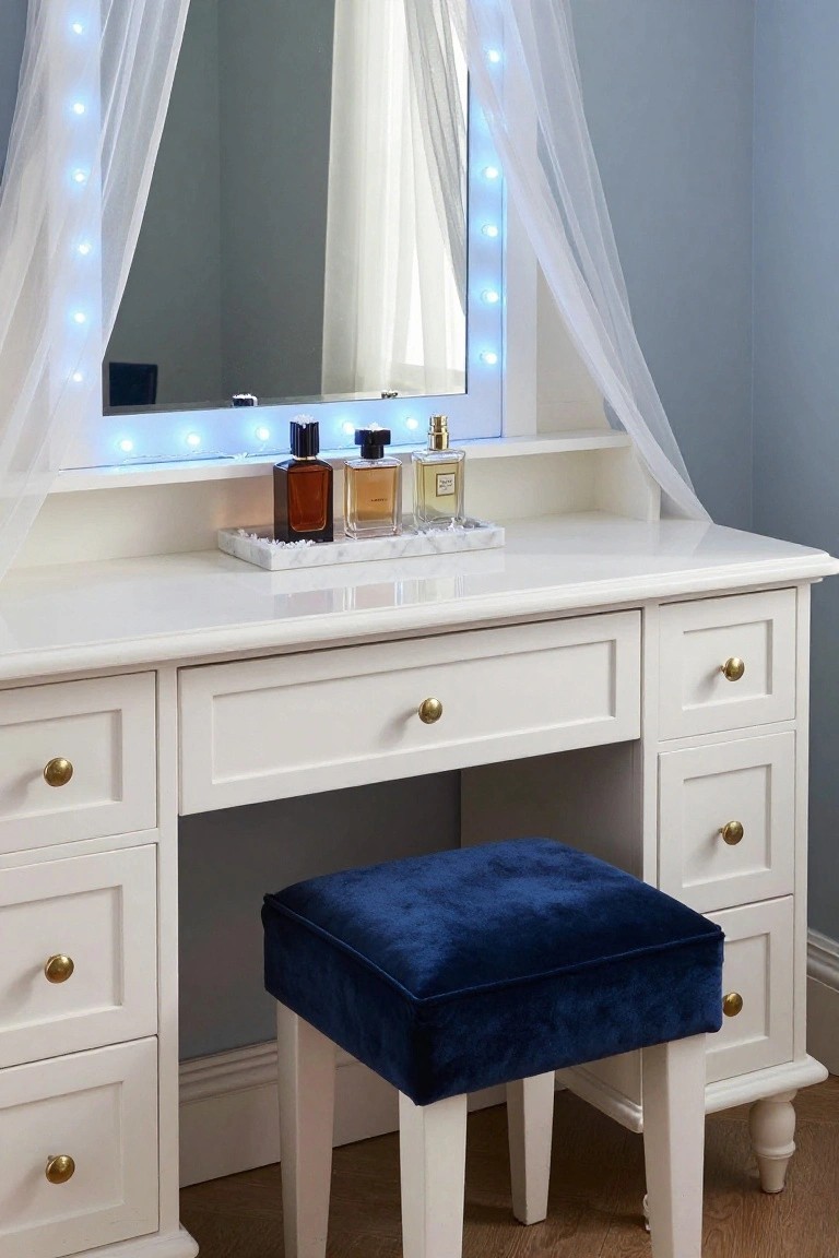 White wooden vanity table with large mirror edged in blue LED lights, perfume bottles on top, marble tray, and blue velvet stool in front, set in a pale blue room with sheer white drapes.