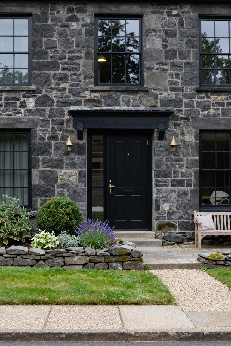 Two-story house with dark gray stone walls, black-framed multi-pane windows, black front door with brass lantern lights, low stone retaining wall with shrubs and perennials, wooden bench, and gravel path to sidewalk.