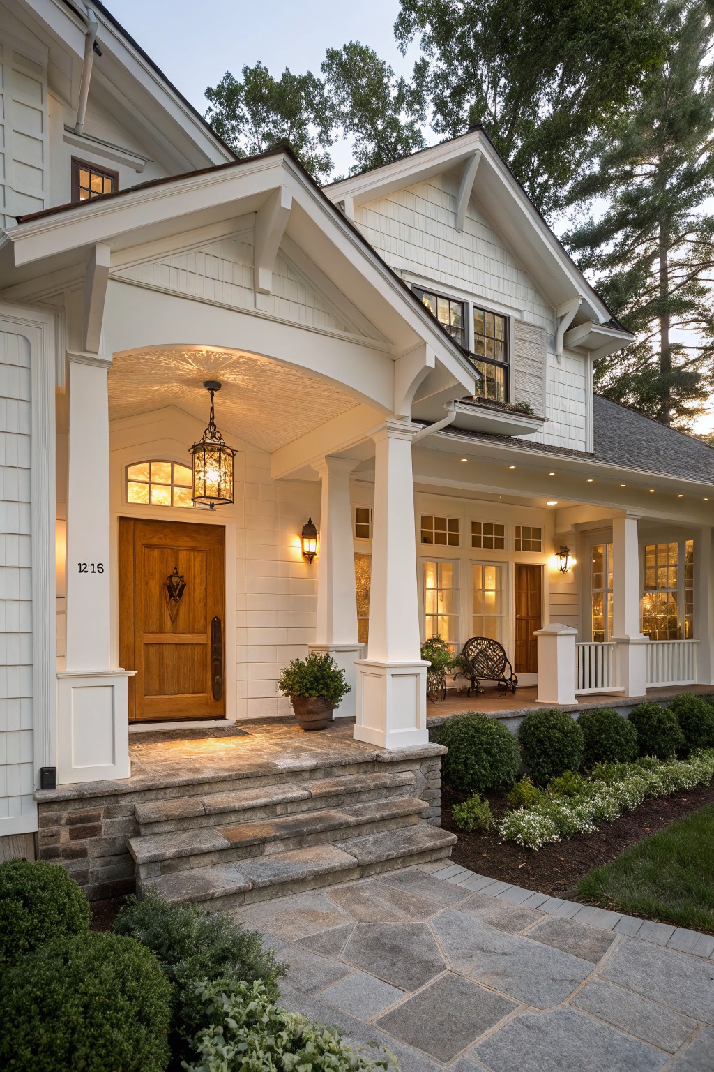 White clapboard house exterior at dusk with covered porch, wooden front door numbered 1216, hanging lantern overhead, wall sconces, columns, stone steps, bluestone path, and boxwood shrubs.