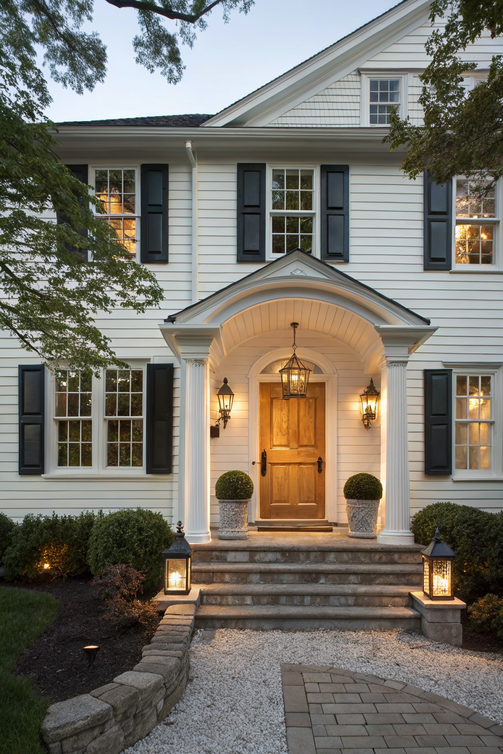 White two-story clapboard house with black shutters, arched covered porch, wood front door, and lantern lights illuminating the entry at dusk, with topiary plants, stone steps, and gravel path.