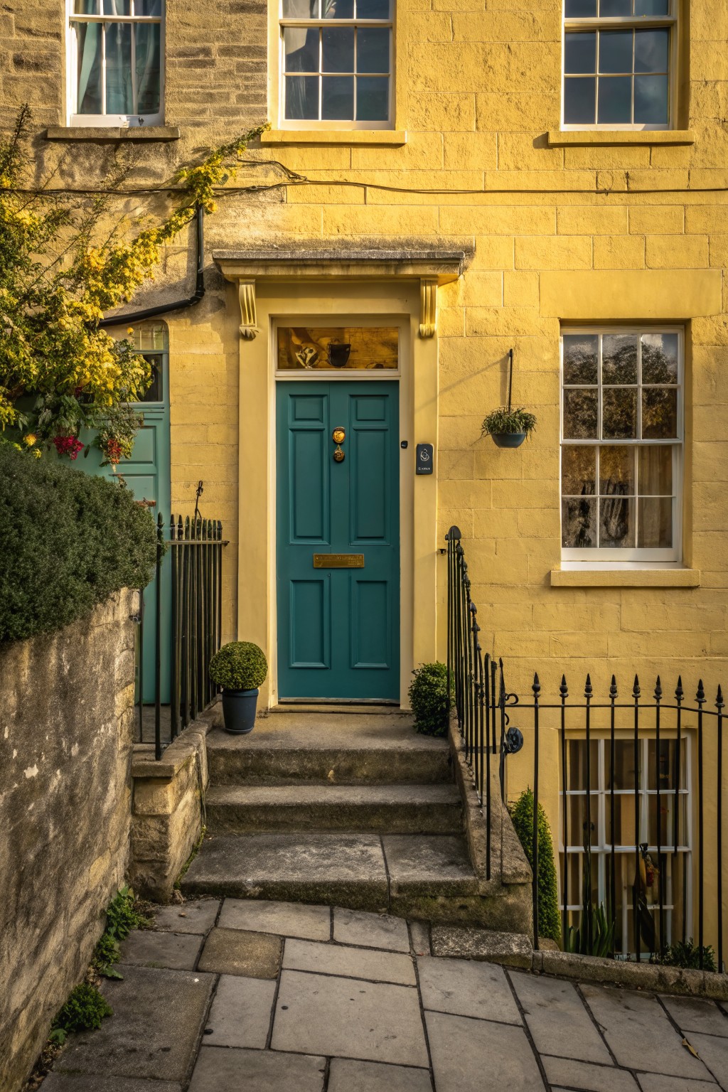 Yellow sandstone terraced houses with teal front doors, black wrought iron railings and gates, stone steps, potted plants, and a hanging basket beside a paved pathway.