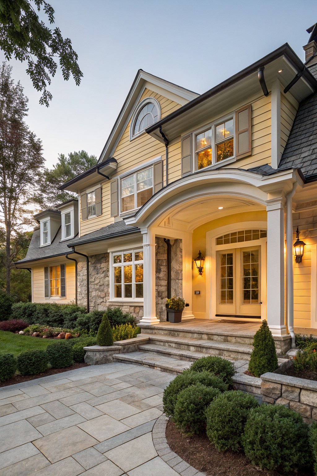 Two-story yellow clapboard house with dark shingled roof, arched entry porch on stone-accented base, white columns, lantern lights, steps, and landscaped shrubs in front yard at dusk.