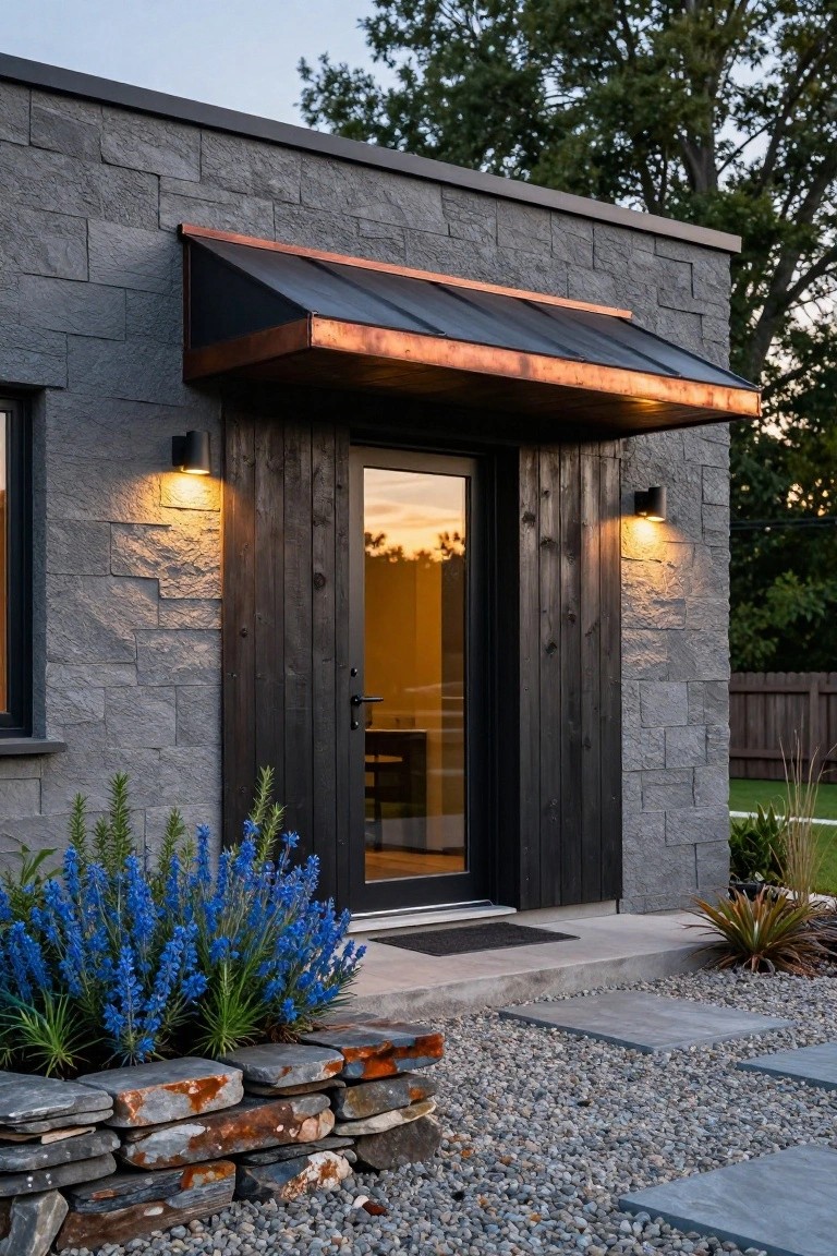 House entrance with gray stone walls, dark wood door with glass panel, copper awning, black wall sconces, blue flowering plants in rock-edged bed, gravel path, and concrete steps.