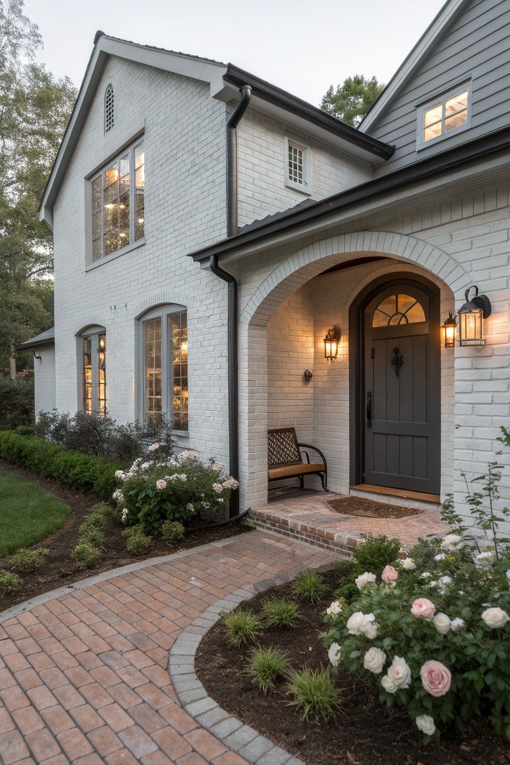 White brick house exterior featuring an arched entry porch with a dark paneled door, wall lanterns, a bench, brick steps and pathway, and surrounding landscaping with shrubs and roses.