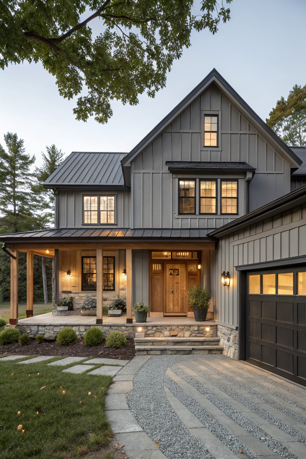 A two-story gray board-and-batten house with gabled roof, standing seam metal roofing, covered front porch with wood posts, black-framed windows, wooden entry door, attached garage, stone foundation, gravel path, and surrounding trees and shrubs at dusk.