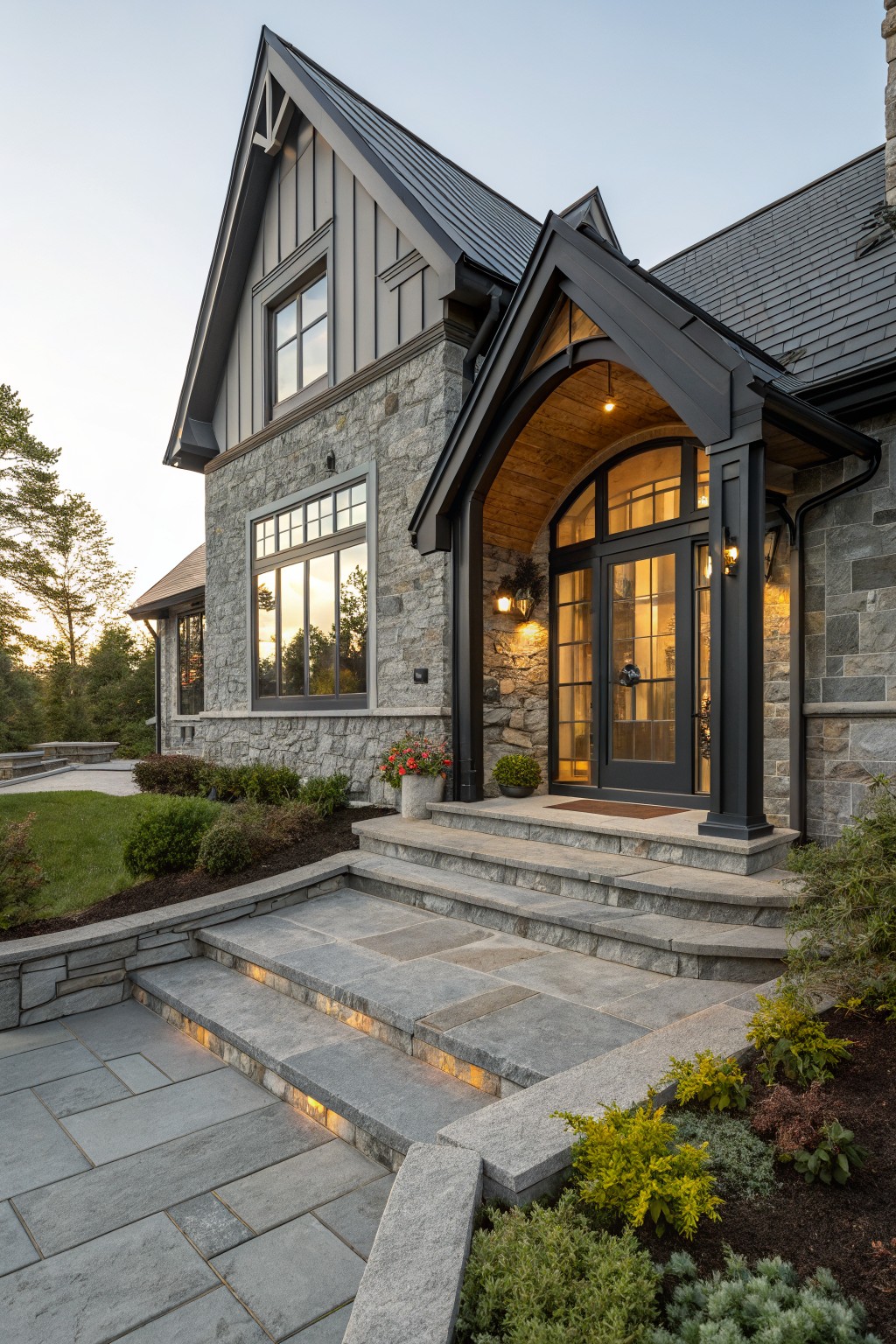 Gray stone house exterior with wood-beamed arched portico over black-framed glass entry doors, stone steps with lighting, and surrounding shrubs and plants.