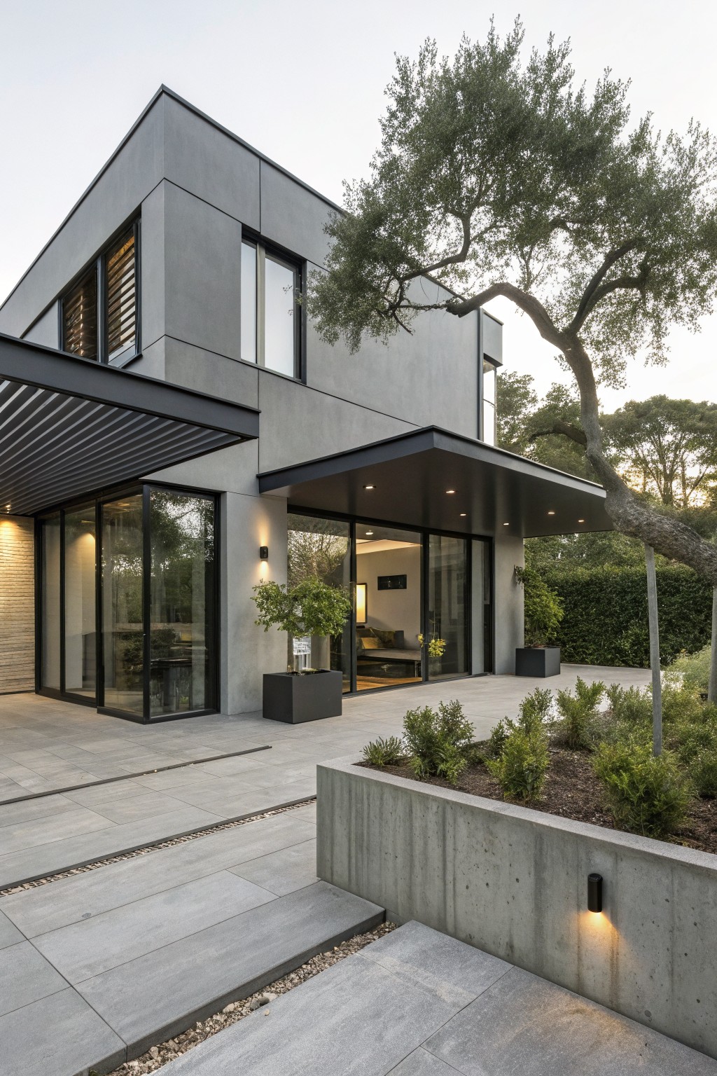 Modern gray concrete house exterior with black metal roof overhang above a large glass entry door and windows, concrete retaining wall with plants, potted trees, and an olive tree nearby.