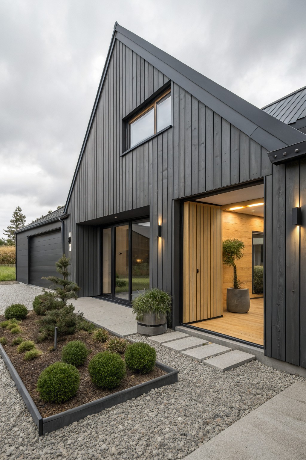 Modern house exterior featuring dark gray vertical wood cladding on a steep gabled roof with black metal roofing, attached gray garage, open blond wood entry door flanked by black lights, gravel pathway, and planted border with shrubs and trees.