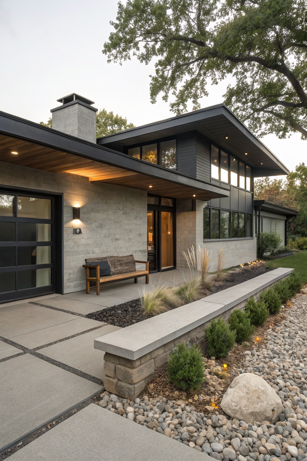 Modern two-story house exterior with light gray brick lower walls, black vertical cladding and metal roof on upper section, wood soffit overhang above entry door and glass garage door, bench on porch, and landscaped bed with ornamental grasses, boxwoods, rocks, and path leading to driveway.