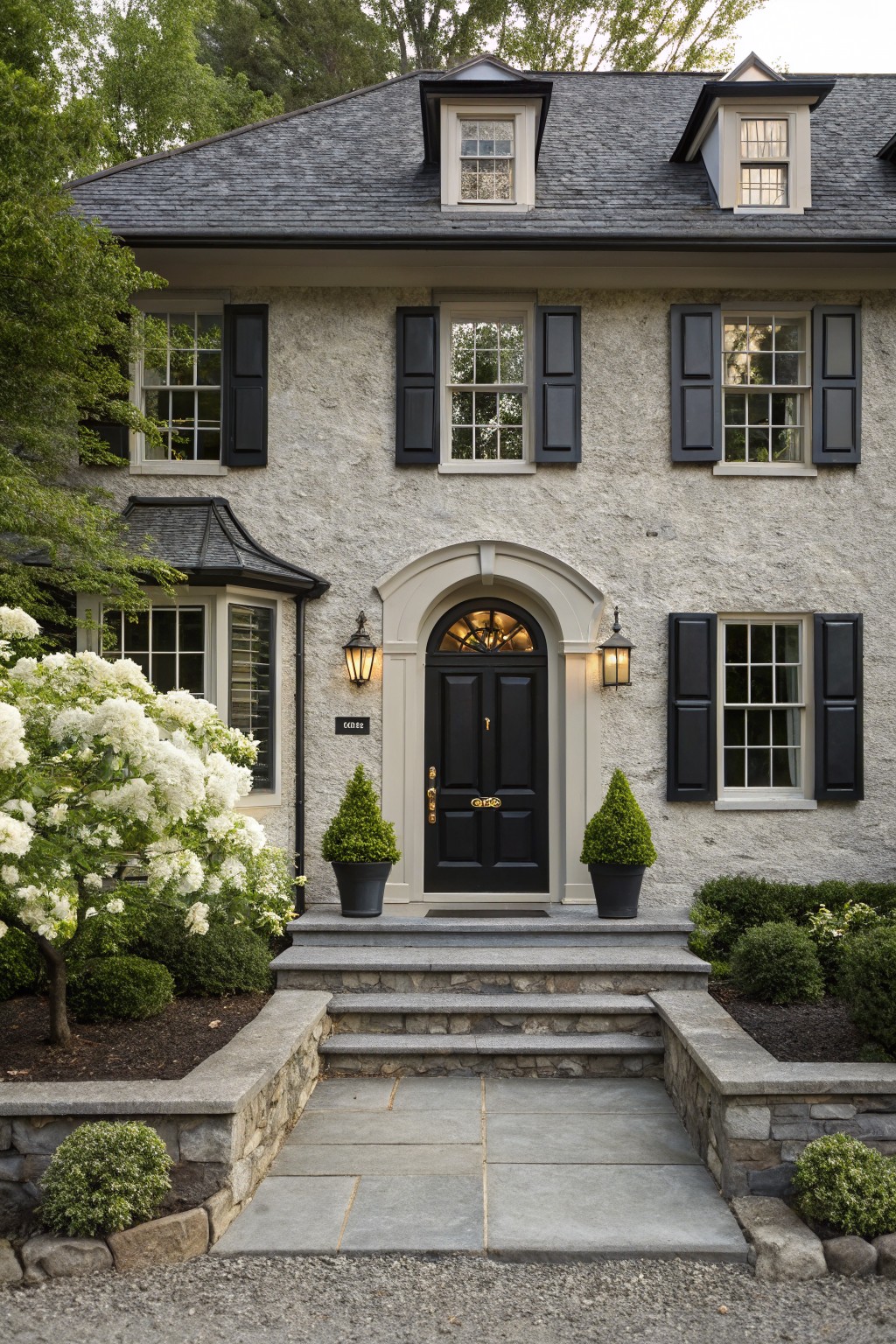 Two-story house with light gray stucco walls, black slate roof, black shutters on multi-pane windows, arched black front door with lanterns, flanked by white flowering shrubs and boxwood topiaries on stone steps leading to gravel driveway.