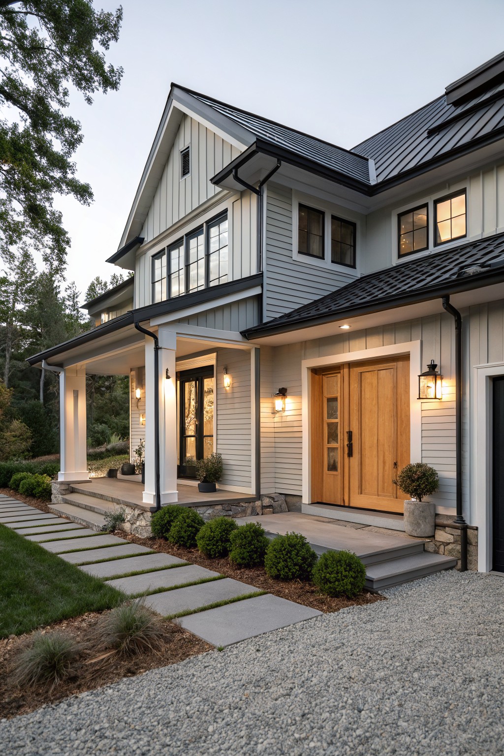 Two-story house exterior featuring light gray shiplap siding, black standing seam metal roof, black trim and gutters, covered front porch with wood double doors, stone accents, and gravel path with shrubs.