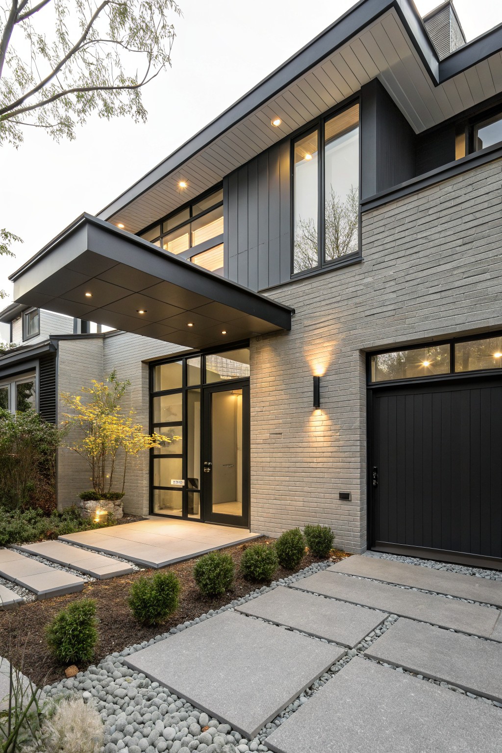 Side view of a modern two-story house with light gray brick facade, black metal cladding and overhanging entry canopy with recessed lights, large glass entry door, black garage door, concrete paver steps and pathway with gravel mulch and low shrubs.