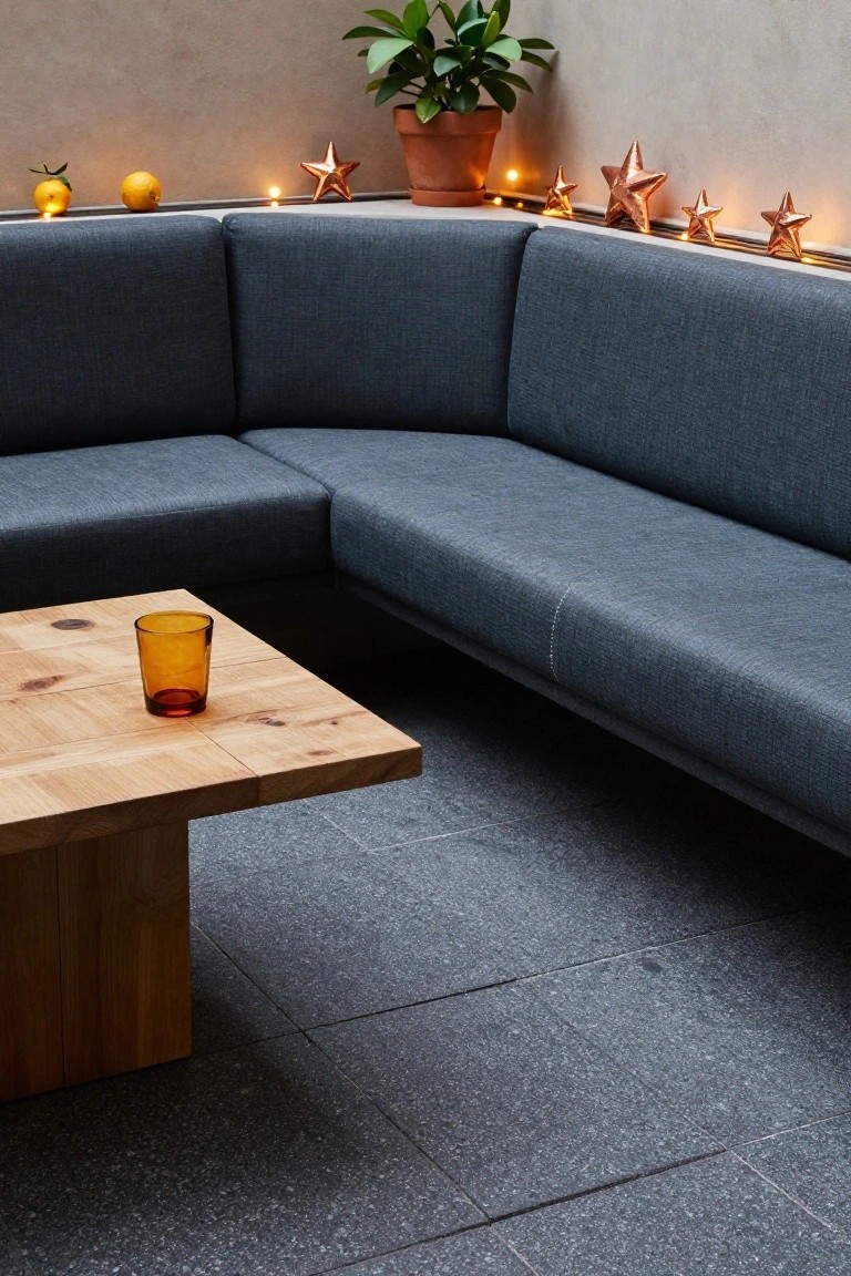 Blue L-shaped sofa against a beige wall ledge decorated with oranges, lemons, copper star ornaments, fairy lights, and a potted plant, with a wooden coffee table and amber glass tumbler on dark tiled floor.