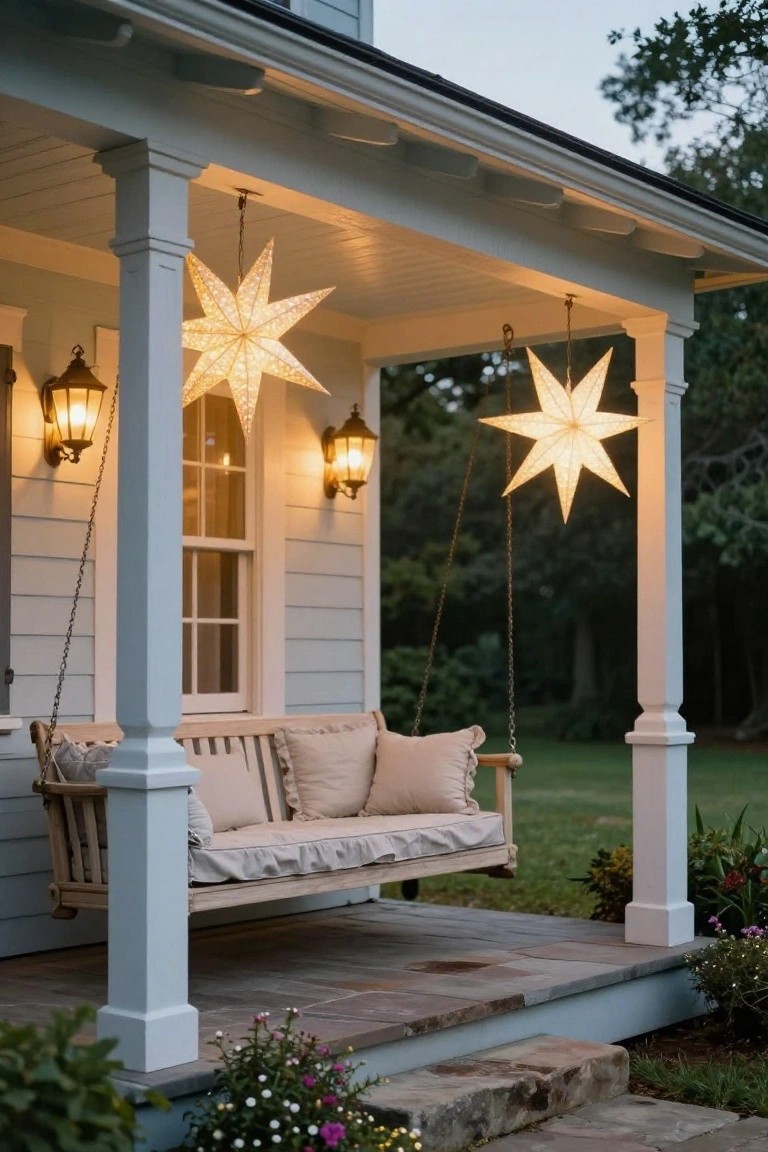 Light blue house porch at dusk with wooden swing, two large white paper star lanterns hanging from ceiling chains between posts, wall lanterns lit, potted plants nearby, and stone steps leading down to yard.