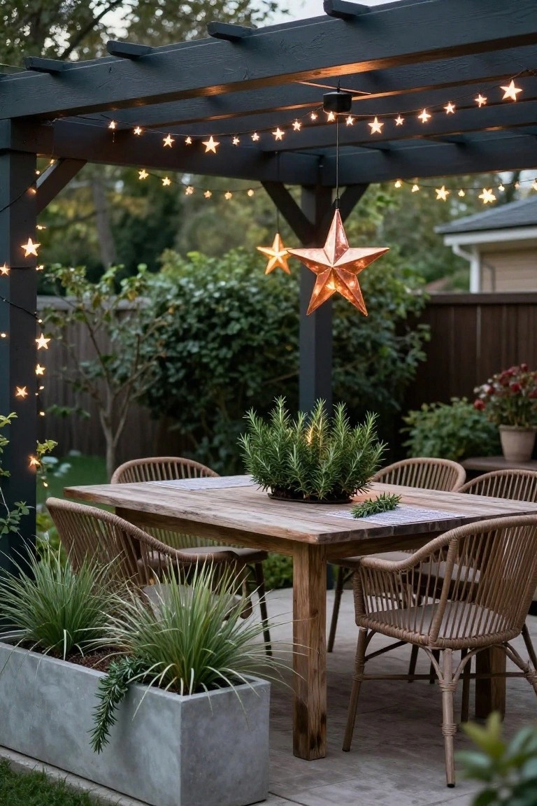 Wooden dining table with rattan chairs and rosemary plant centerpiece on a patio under black pergola strung with star lights and a copper star pendant hanging above, potted grasses on sides.