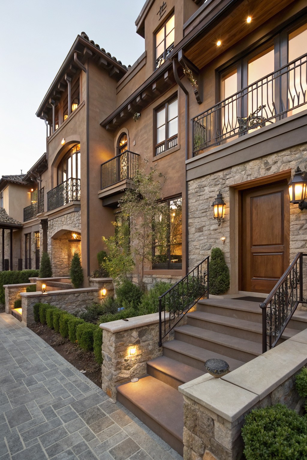 Two-story house exterior with textured brown stucco walls, gray stone base and entry surround, wooden double front door, wrought-iron railings on concrete steps, landscape lights, boxed shrubs, and paver walkway.