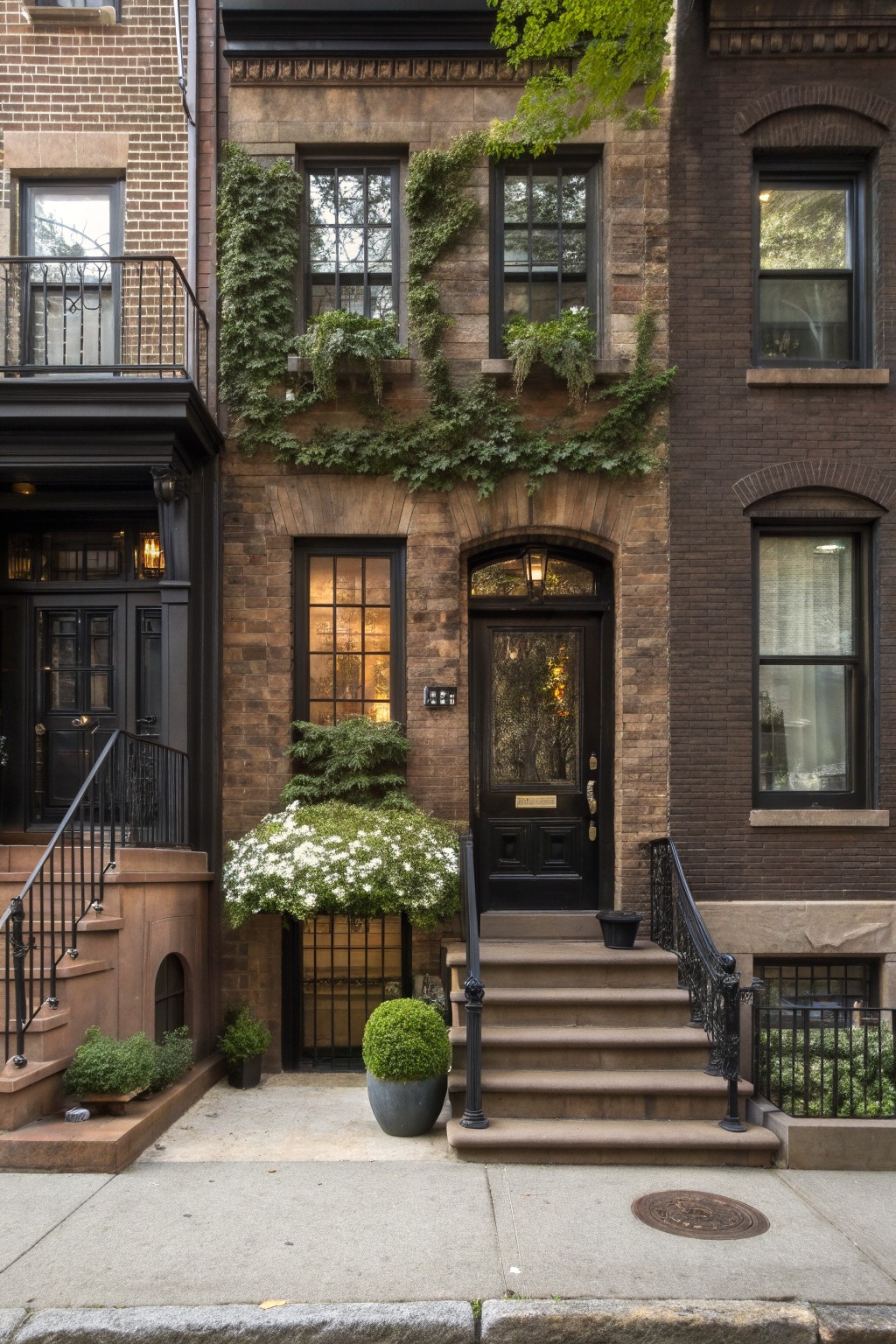Row of three-story brown brick townhouses on a city sidewalk, central house with climbing ivy on facade, black front door, potted plants on stone steps, and greenery in window boxes.