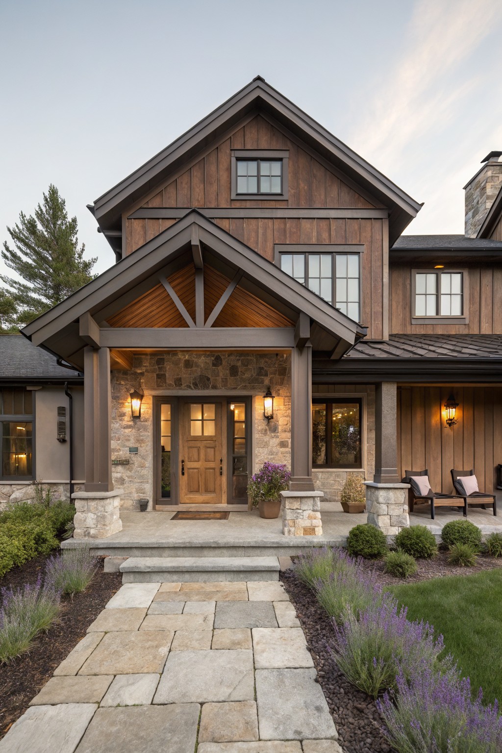 Front view of a two-story house with brown vertical wood siding, covered porch supported by stone pillars, double wooden doors flanked by lanterns, and a stone path with lavender borders leading to the entry.