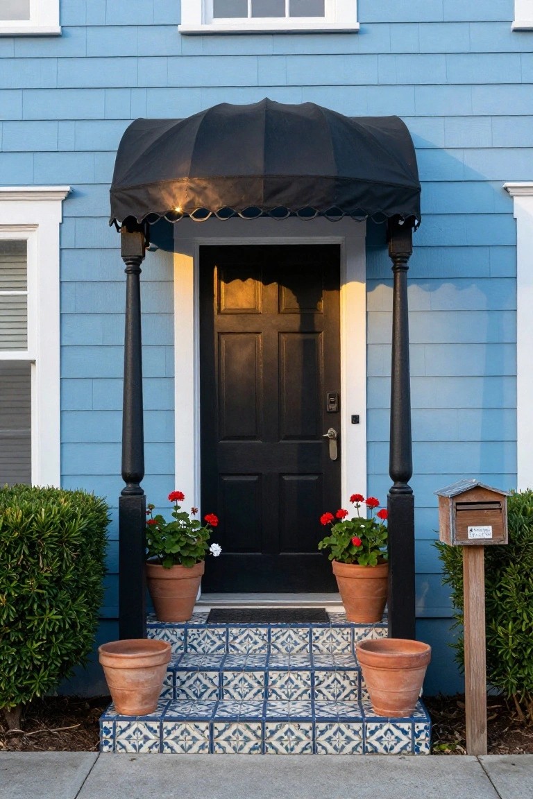 Front exterior of a light blue shingle-style house with a black curved awning over a black paneled door, flanked by black columns, red geraniums in terracotta pots on either side, blue tiled steps, and a wooden mailbox post.