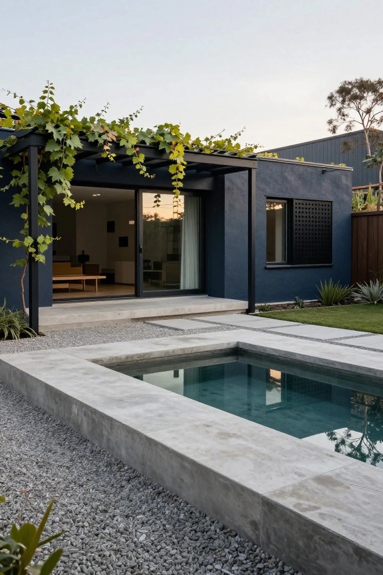 Dark navy blue rendered house exterior with black-framed sliding glass doors and window, vine-covered metal pergola extending over a concrete terrace adjacent to a small rectangular pool edged in concrete and surrounded by gravel ground cover.