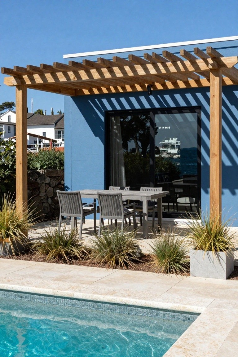 Wooden pergola shading a grey rectangular dining table with six matching chairs on a beige stone patio next to a blue house wall with glass door and an in-ground swimming pool, with grasses and plants in planters nearby.