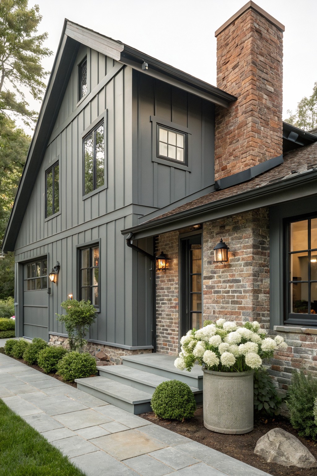 Side exterior of a two-story gray board-and-batten house featuring a brick chimney, brick entry base with door and windows, attached garage, stone pathway, potted white hydrangea, boxwoods, lanterns, and surrounding lawn and trees.