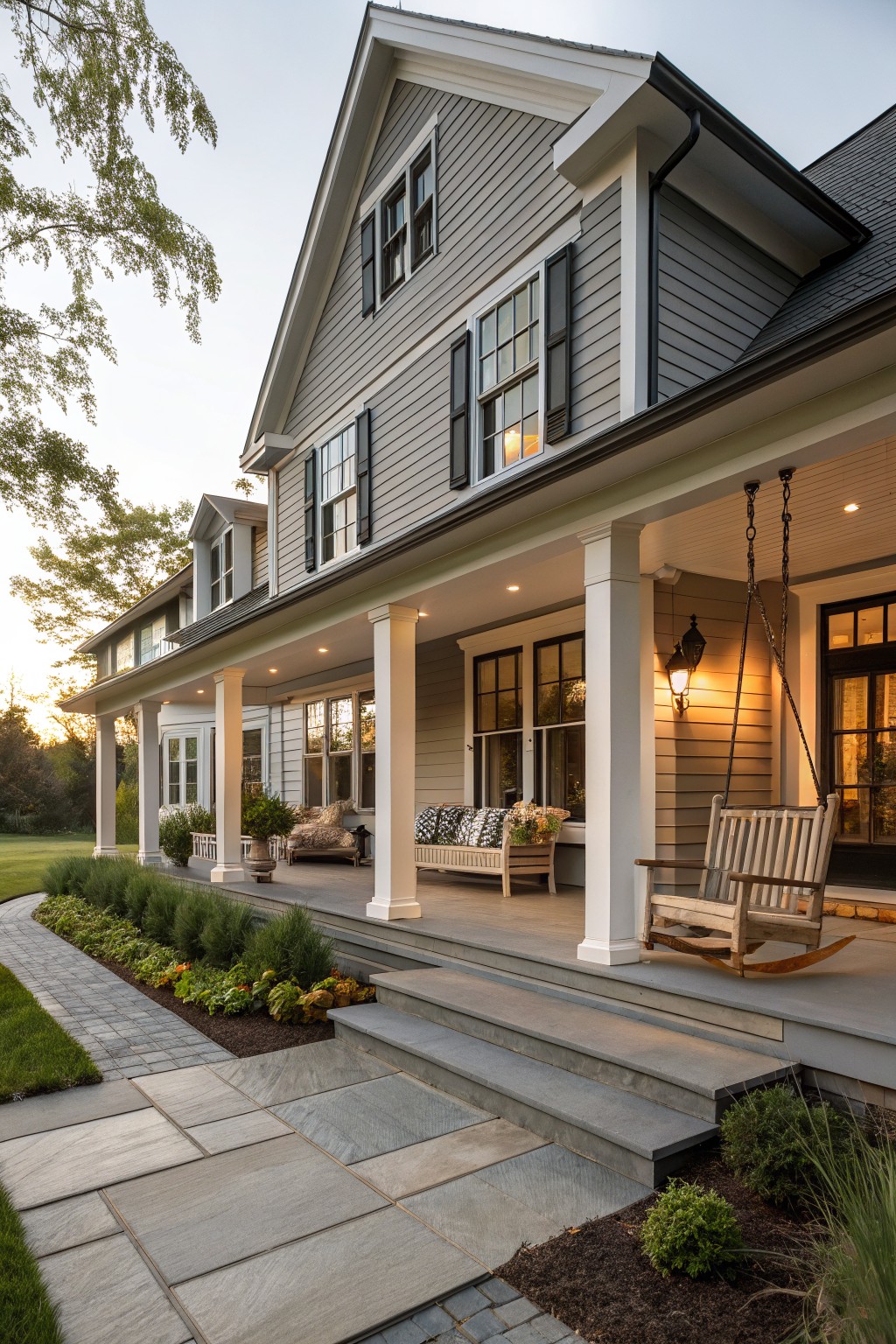 Gray shingle-sided two-story farmhouse with wraparound covered porch supported by white columns, wooden porch swing and rocker chair, steps to bluestone path with low plantings and lawn at dusk.