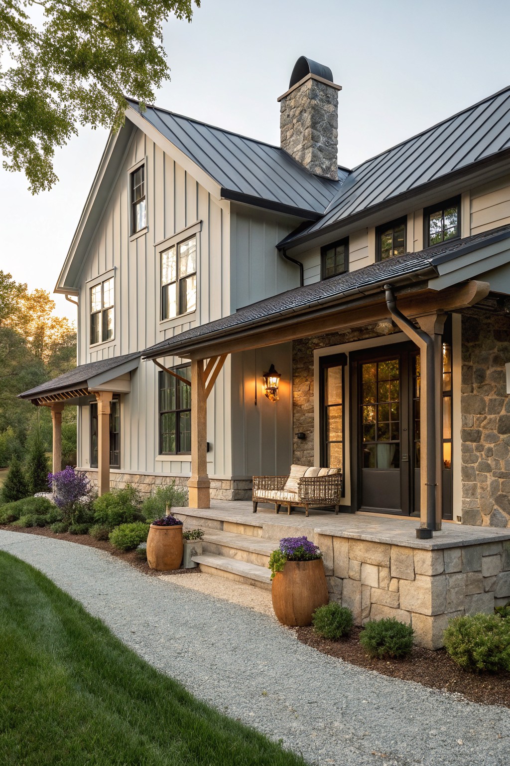 Two-story farmhouse exterior with light gray board-and-batten siding, dark standing-seam metal roof, stone chimney and porch foundation, covered front porch with wood posts and bench seating, gravel path, and low plantings.