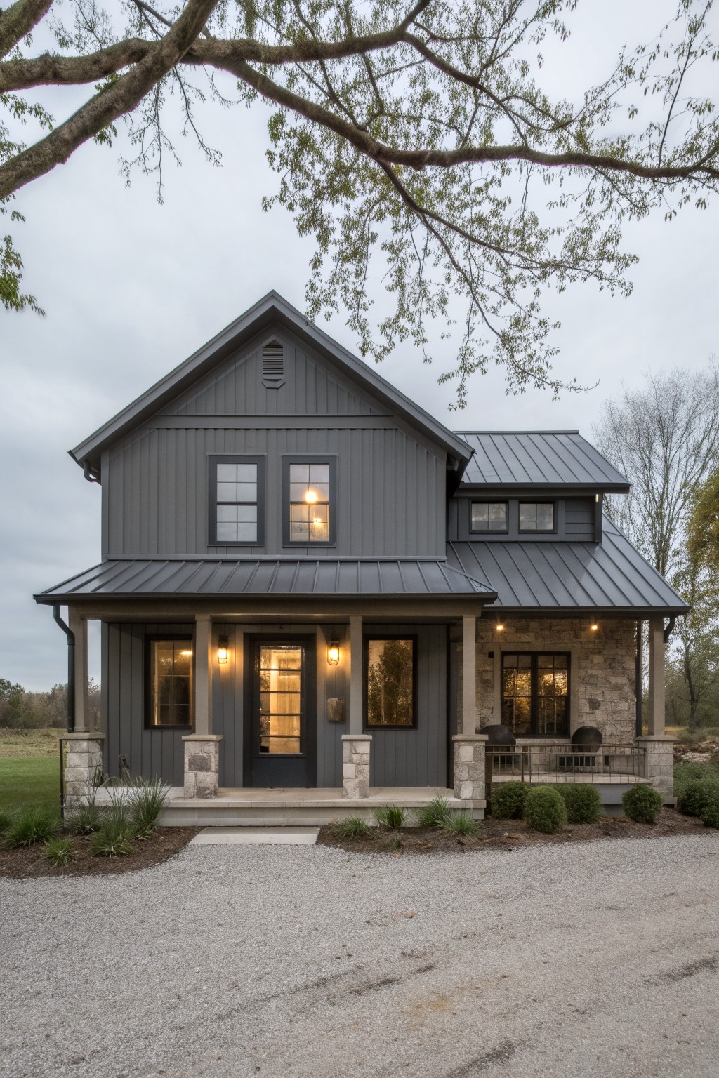 Gray board-and-batten sided farmhouse with standing seam metal roof extending over the front porch, stone columns, lanterns, and gravel driveway in front of the house.