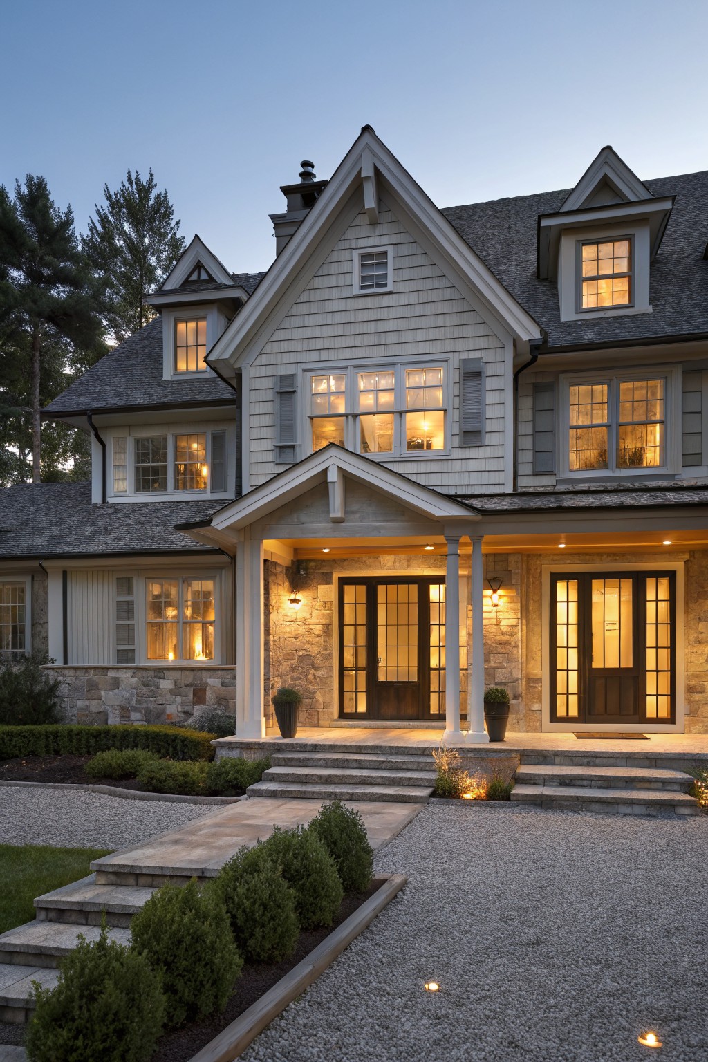 Two-story gray shingled house with gabled roof, covered front porch supported by stone columns, double dark wood doors, lit windows, steps leading to gravel driveway edged by boxwood shrubs at dusk.