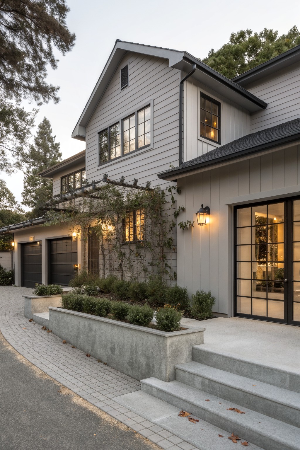 Side exterior of a two-story gray board-and-batten house with black garage doors, vine-covered metal trellis, black-framed glass entry doors, concrete steps and raised planters along a curved driveway edged in pavers.