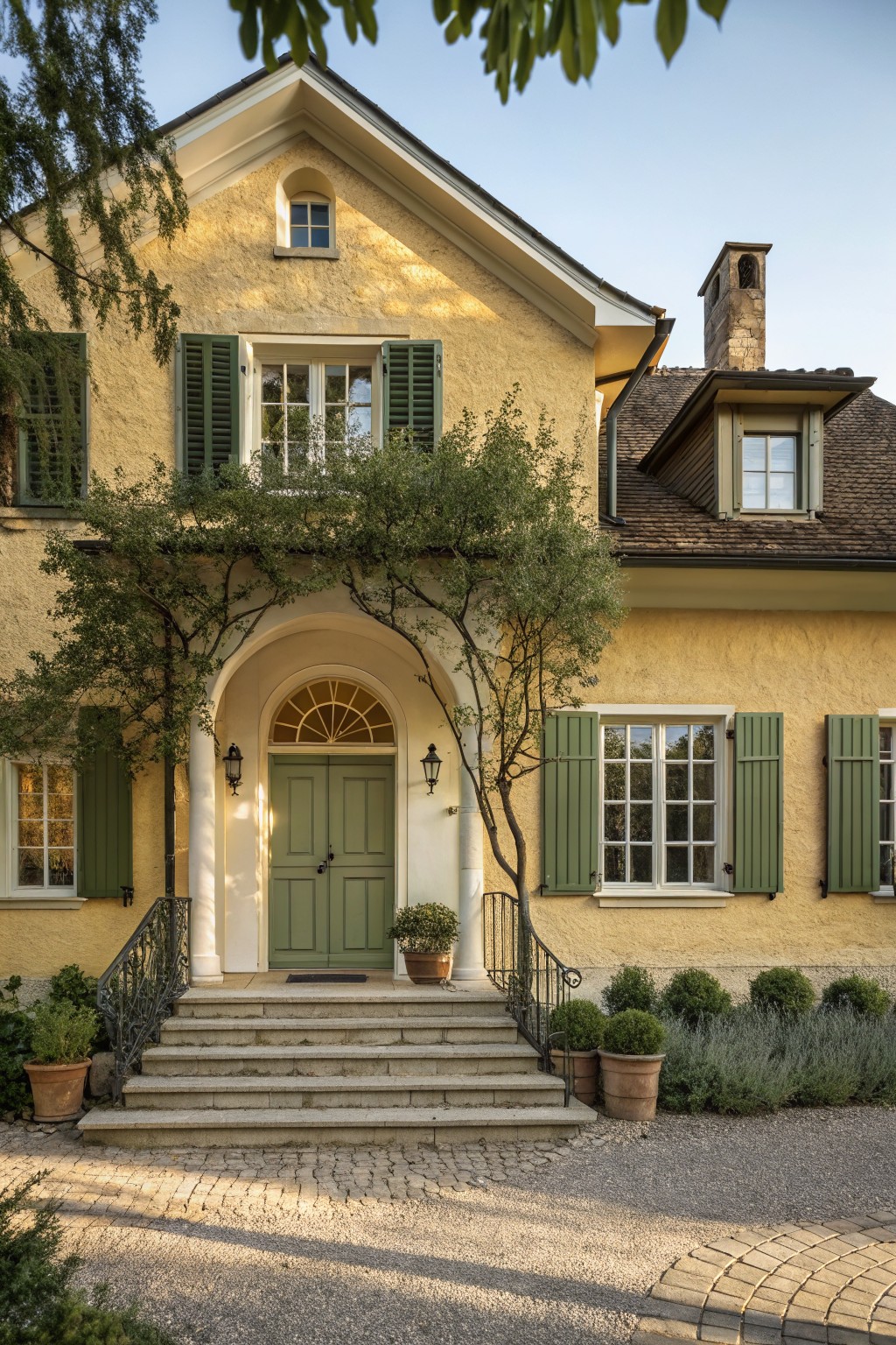 Yellow stucco cottage exterior with arched green double doors, olive tree branches overhead, green shutters on windows, stone steps, wrought iron railing, potted plants, and gravel path.