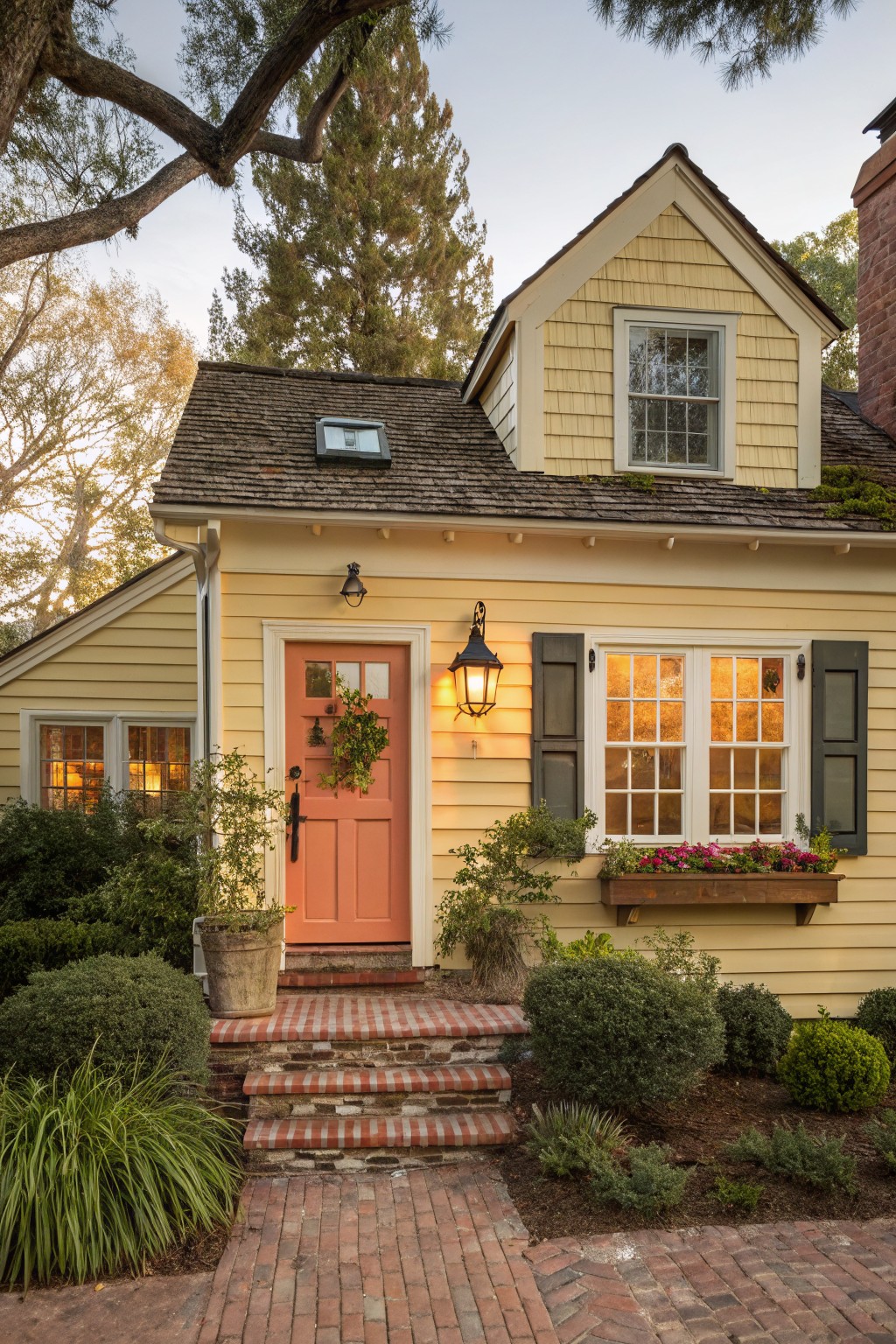 Pale yellow shingle-style cottage with coral paneled front door, green shutters on windows, flower-filled window box, hanging lanterns, brick entry steps, potted plant, shrubs, and trees in golden hour light.