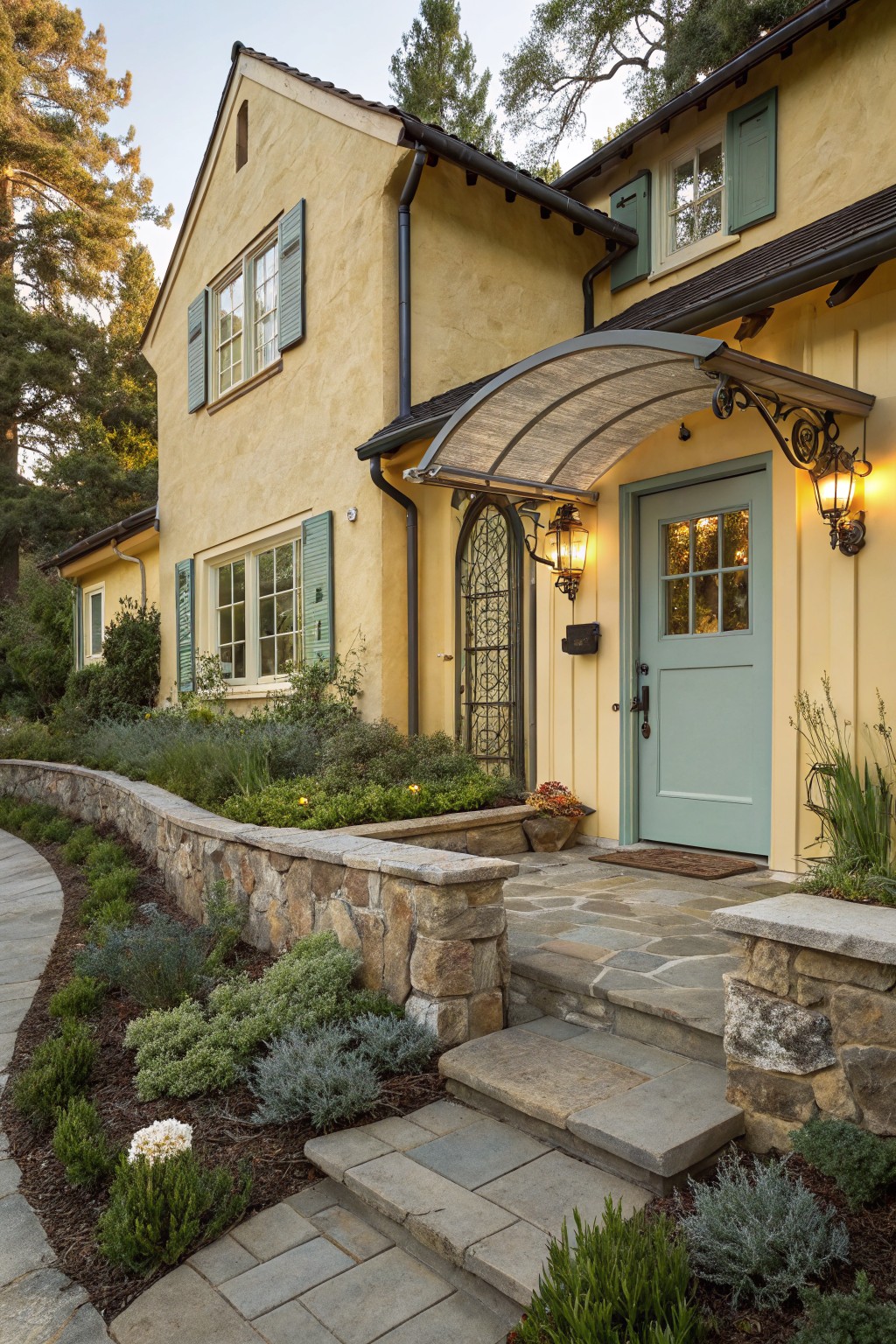 Yellow stucco house exterior featuring a teal paneled front door under a curved metal awning, with green shutters on windows, wrought iron lanterns, stone retaining walls, steps, and low groundcover plants.
