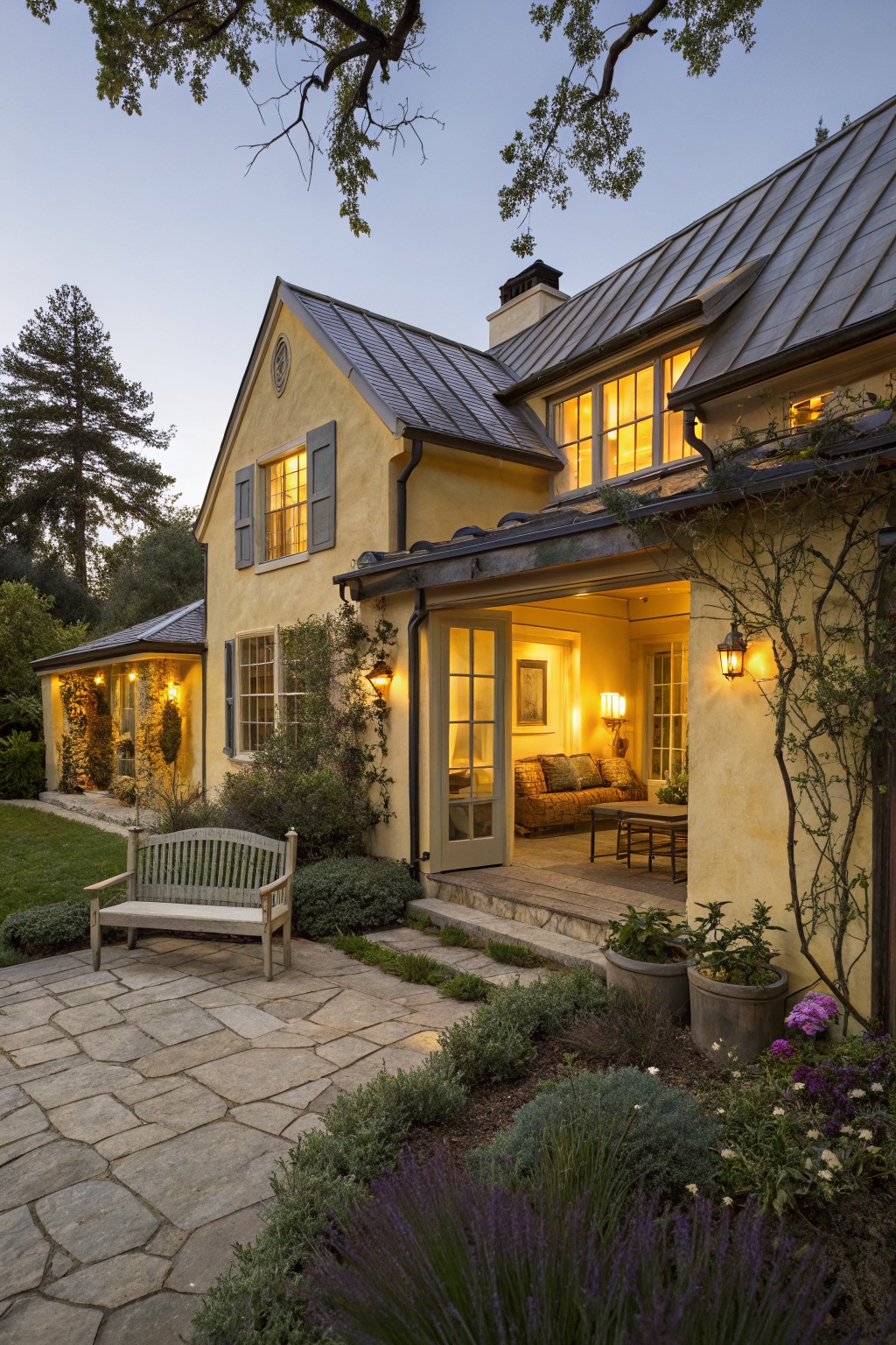 Evening photo of a two-story yellow stucco cottage with dark metal roof, lit windows with shutters, open French doors to a covered patio with sofa, wooden garden bench on stone path, and surrounding plants and trees.
