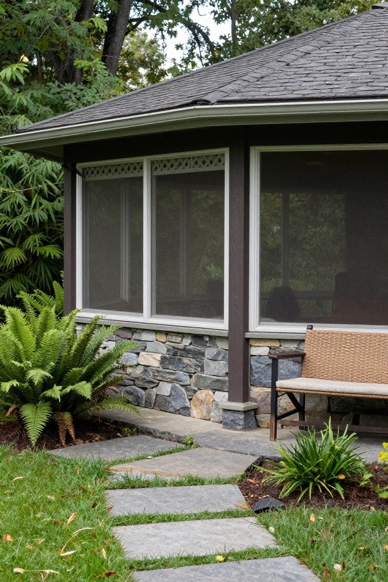 Craftsman-style screened porch with three large screened windows topped by decorative lattice, dark wood posts and frame, stone base, wicker bench nearby, ferns and plants at the base, stone path leading from grass lawn surrounded by trees.