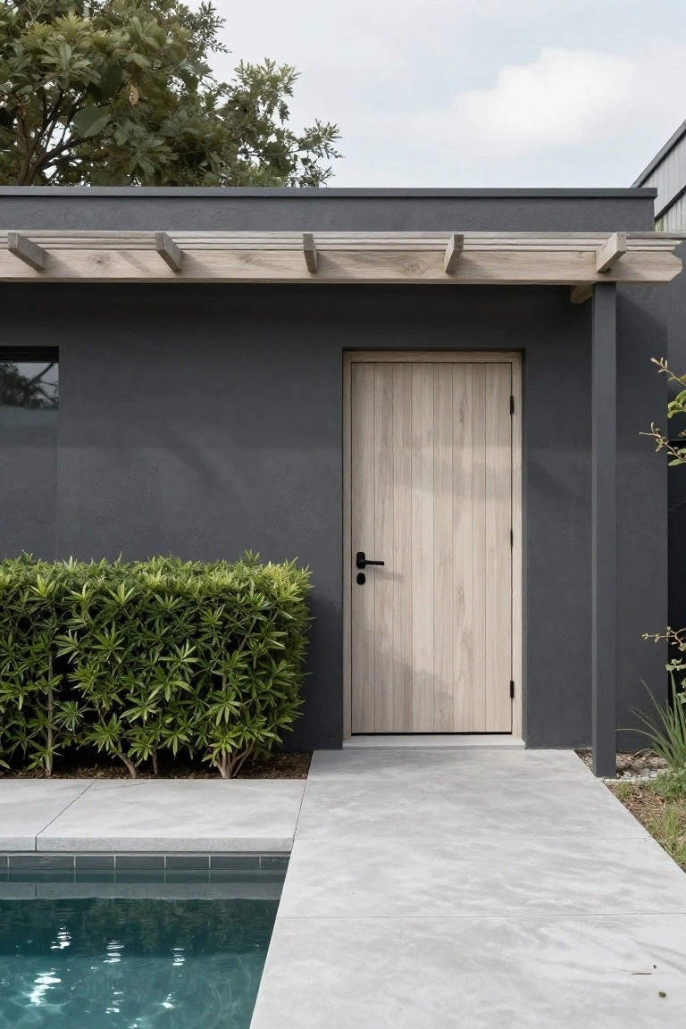 Dark gray stucco house wall with light vertical wood door, black handle, wooden pergola above, green boxwood hedge beside, gray concrete path to tiled pool edge.